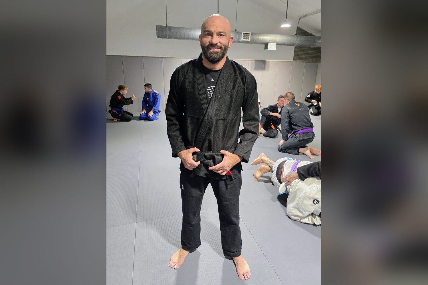 Josh Smith in a black gi wearing his new Brazilian Jiu Jitsu black belt on the mats at Haven on the Hill in Syracuse